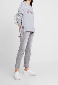 Grå sweatshirt med broderad text, sidsprund, i kombination med ljusgrå jeans och vita platåsnickers. Mot en neutral bakgrund.