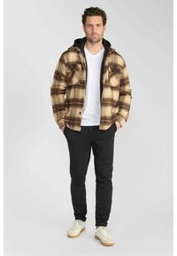 Chemise en flanelle à carreaux beige et marron, avec une capuche noire, deux poches avant, associée à un pantalon noir et des baskets blanches.