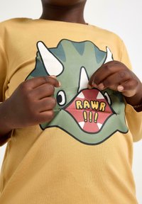 Gul bomulds T-shirt med et grafisk motiv af et grønt dinosauransigt med hvide tænder og overdrevne træk, sammen med teksten "RAWR!!!" i sort.
