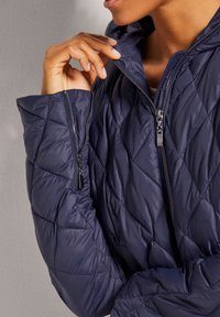 Marineblaue, wattierte Pufferjacke mit hohem Kragen und doppelt verstellbarem Reißverschluss. Verfügt über eine glänzende, glatte Textur und einen taillierten Schnitt.