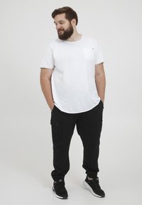 Weißes T-Shirt mit Rundhalsausschnitt und Brusttasche, kombiniert mit schwarzen Cargo-Hosen mit Seitentaschen und elastischen Bündchen, abgerundet mit schwarzen Sneakers.