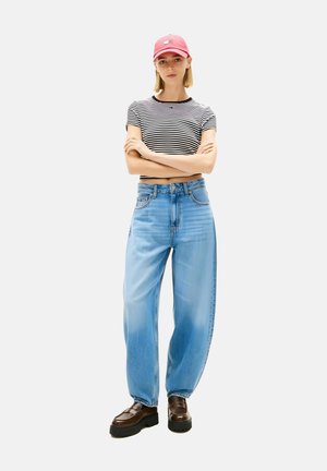 Giovane in piedi con le braccia incrociate, indossa un berretto rosa, una maglietta corta a righe bianche e nere, jeans larghi azzurri e scarpe con zeppa marroni.