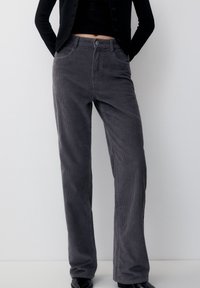 Pantalon en velours côtelé gris avec une coupe droite, présentant une texture en côtes verticales, des poches avant et une fermeture à bouton.