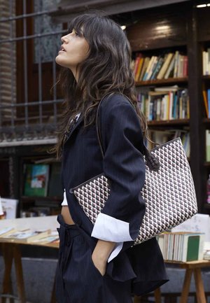 Zwarte pinstripe blazer, witte mouwen van een shirt, houdt een patronen tote bag vast met geometrisch ontwerp in maroon, beige en bruine tinten. Achtergrond van een boekwinkel.