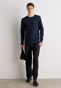 Camisa de manga larga en color navy con un pequeño logo en el pecho, combinada con pantalones negros. El modelo sostiene un bolso negro y lleva unos zapatos negros.