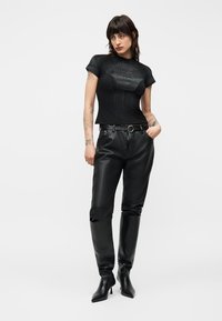 Moteris su trumpais tamsiais plaukais, dėvinti aptemptą juodą tekstūruotą trumpų rankovių palaidinę, juodas odines kelnes ir juodus ilgaaulnius aukštakulnius batus iki kelių.