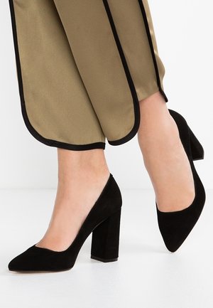 Jambes portant un pantalon beige avec un liseré noir et des escarpins en daim noir à talons blocs sur un sol blanc.