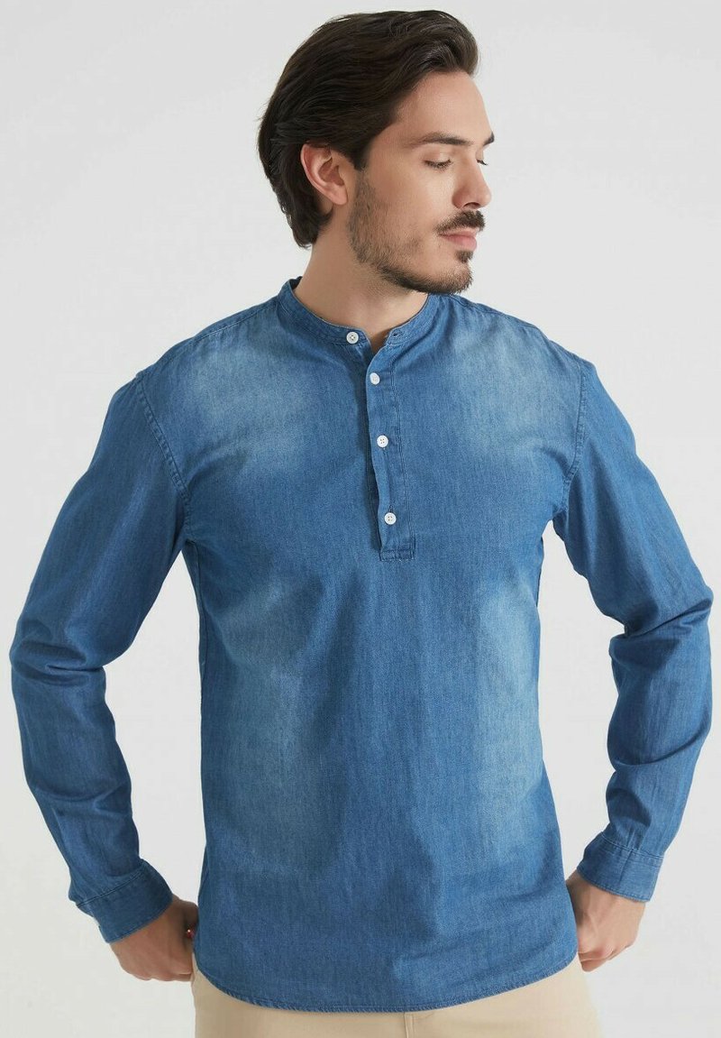 Homme aux cheveux bruns portant une chemise en denim bleue à manches longues avec des boutons, ajustant l'ourlet de sa chemise, devant un fond clair et uni.