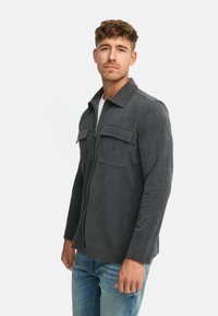 Grå fløjlsk jacket med lynlås foran, to brystlommer, krave og lange ærmer, iført over en hvid skjorte og blå jeans.