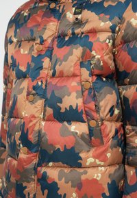 Doudoune dans un motif camouflage multicolore avec des teintes rouges, brunes et bleues. Comprend un tissu matelassé et des boutons-pression bruns.