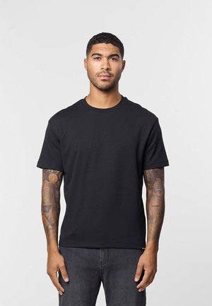 Jeune homme aux cheveux courts et tatouages sur les deux avant-bras, portant un t-shirt noir uni et un jean foncé, debout devant un fond clair.