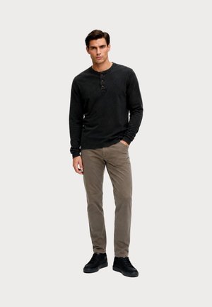 Mand iført en sort langærmet henley-skjorte, brune bukser og sorte sneakers står med den ene hånd i lommen på en ensfarvet baggrund.