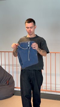 Vyras su tatuiruotėmis laiko mėlynę gobtuvinę viduje, šalia rudos pupelių kėdės ir oranžinės turėklos.