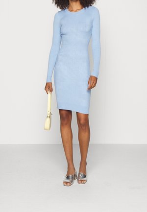 Femme portant une robe longue à manches longues en tricot côtelé bleu clair, tenant un petit sac à main crème, et portant des talons blocs argentés ouverts à l'avant.