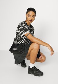 Black graphic t-shirt with white text, dark skirt, and black sneakers. Small black shoulder bag with a logo. Model in a crouched pose.