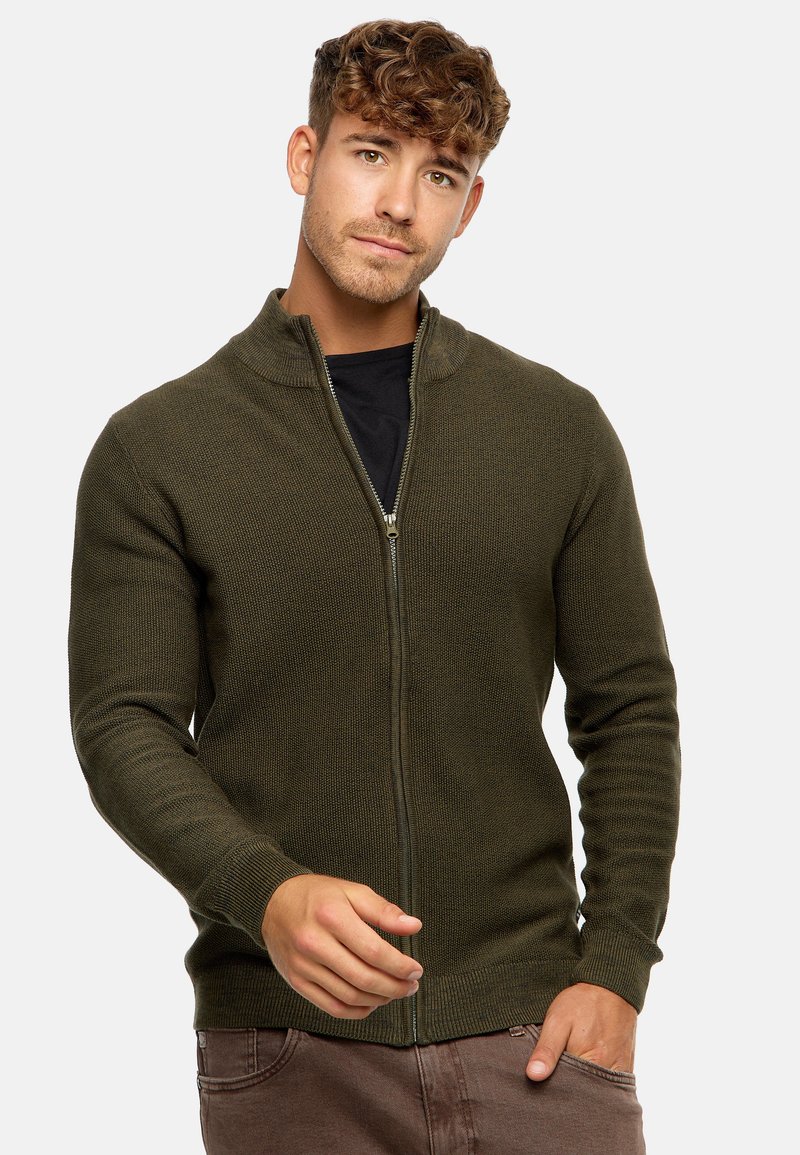 Sudadera verde oliva con cremallera, de diseño de punto texturizado, con puños y dobladillo acanalados, y un cuello alto. Lleva puesta sobre una camiseta negra.