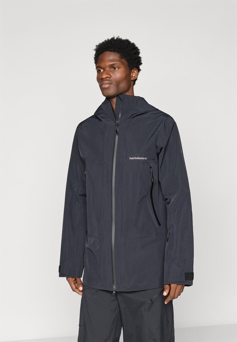 Muž v čiernej nepremokavej bundě s kapucňou a so zipsovými vreckami, ktorý stojí proti jednoduchému pozadiu, v čiernych nohaviciach.