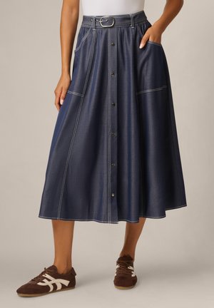 Vrouw draagt een donkerblauwe midi-rok met knopen, riem en zijzakken, gecombineerd met bruine en witte sneakers.