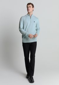 Lichtblauwe ritstrui van gebreide stof, met een staande kraag en geribbelde manchetten, gecombineerd met zwarte broek en zwarte schoenen.