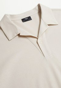 Polo beige à manches courtes avec col plié, une patte de boutonnage avec un bouton, et une étiquette de marque noire à l'intérieur de l'encolure.