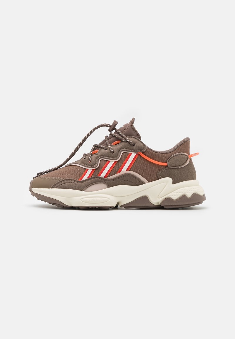 Brown athletic sneakers with red and white stripes, rounded toe, textured fabric, and a chunky white midsole. Features lace closure and accent details.