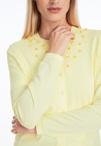 Yellow cardigan featuring a floral design with yellow fabric flowers and beads on the chest, smooth texture, and button closures.
