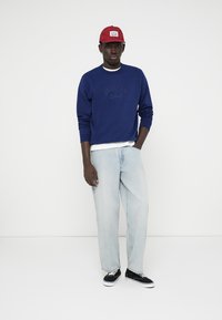 Young man wearing a red cap, navy sweatshirt with text, light blue jeans, and black sneakers standing against a white background.