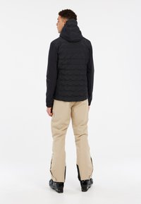 Schwarze Pufferjacke mit Kapuze und strukturierten Einsätzen, kombiniert mit beigen Skihosen mit seitlichen Reißverschlüssen und schwarzen Skistiefeln.