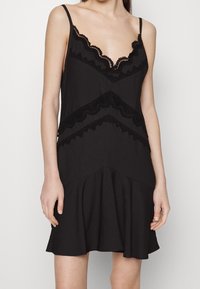 Černé šaty s tenkými ramínky, zdobené krajkovým lemem podél výstřihu a švů, se přiléhavým trupem a rozšířenou sukní. Hladká textura.