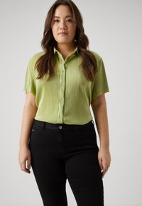 Jauna moteris su ilgais rudais plaukais, vilkinti šviesiai žalią trumpomis rankovėmis mygtukų marškinėlį ir juodas kelnes, stovi prieš paprastą foną.