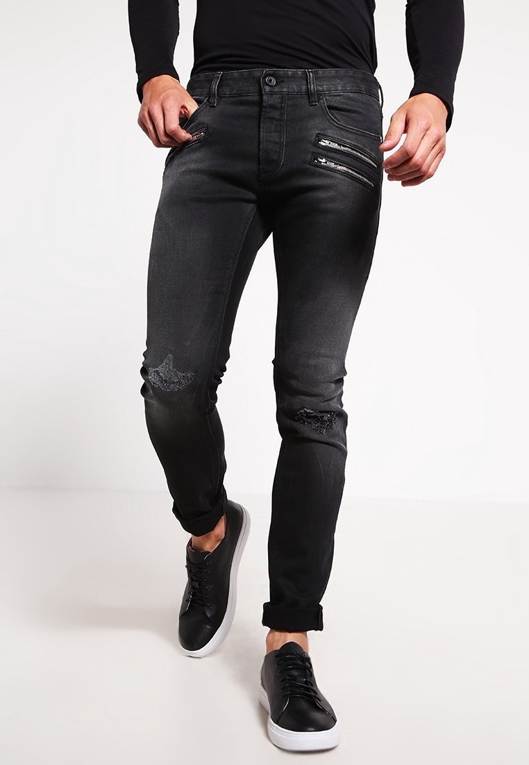 Homme portant un jean slim noir avec des genoux déchirés et des poches zippées, assorti à des baskets noires à semelles blanches.