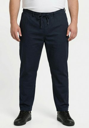 Mann trägt marineblaue Kordelzughose und schwarze Sneakers, steht vor einem schlichten hellen Hintergrund.