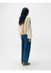 Beige fleece vest over en hvid tekstureret langærmet top, parret med løse blå jeans. Outfitet inkluderer brune fodtøj.