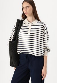 Polo z dolgimi rokavi v črtah v off-white in temno modri barvi s klasičnim ovratnikom. V kombinaciji s temno modrimi hlačami in črno torbo, obešeno čez ramo.