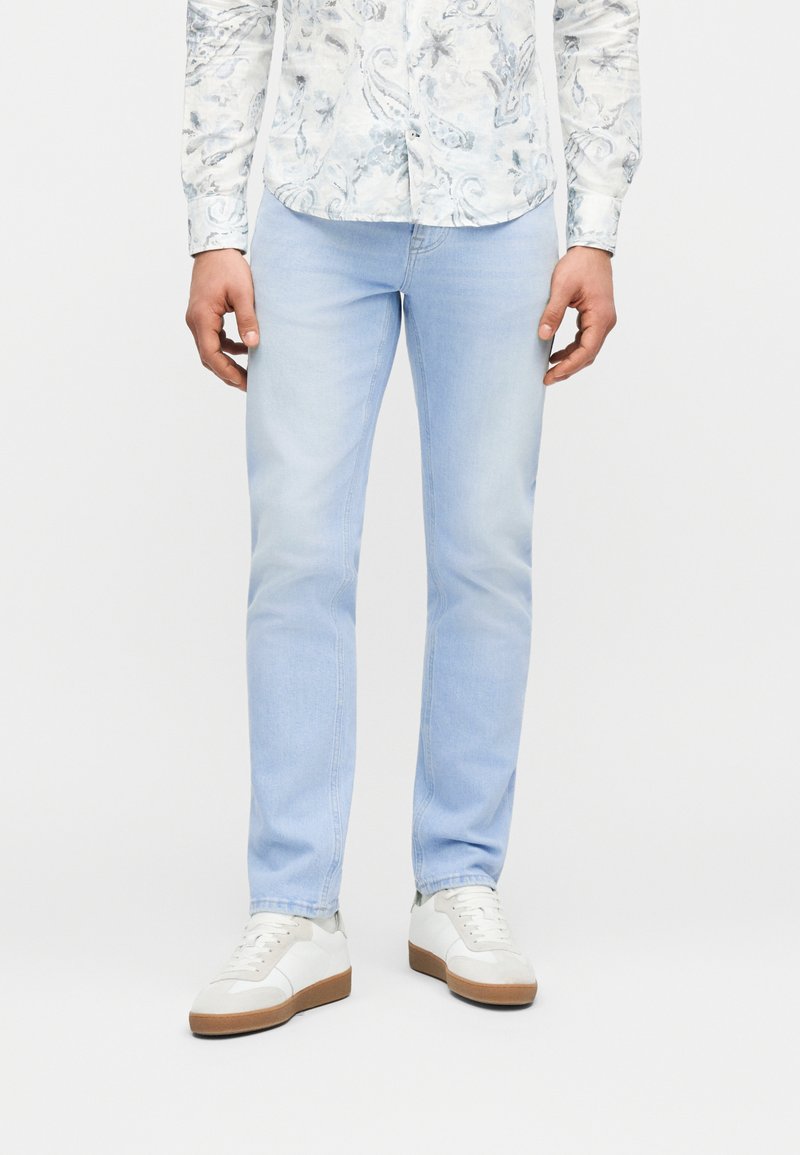 Άνδρας που φοράει ανοιχτό μπλε τζιν slim-fit, λευκό πουκάμισο με λουλουδάτο μοτίβο μακριά μανίκια και λευκά αθλητικά παπούτσια με καφέ σόλες, στέκεται μπροστά σε λευκό φόντο.