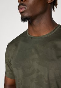 Olive green short-sleeve t-shirt with a subtle camouflage pattern, round neckline, and textured fabric. Close-up view of the neckline.