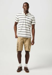 Vit randig polo tröja, khakifärgade cargoshorts med sidorfickor, bruna skor och vita strumpor, stående mot en neutral bakgrund.