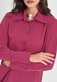 Femme les bras croisés portant une blouse magenta à boutons et un collier avec deux grandes perles blanches.