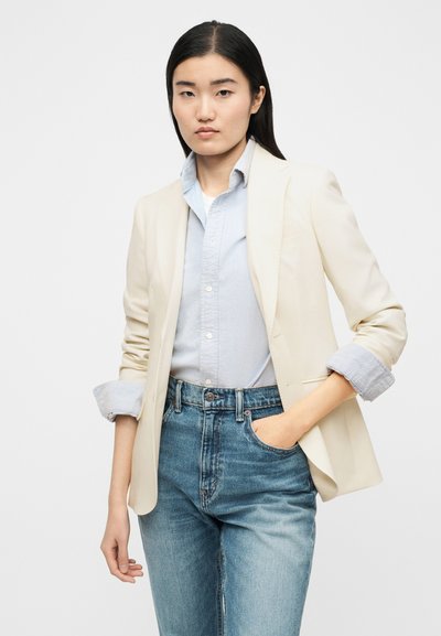 Νεαρή γυναίκα με μακριά σκούρα μαλλιά, ντυμένη με εκρού σακάκι, γαλάζιο πουκάμισο με κουμπιά και μπλε τζιν, στέκεται με το ένα χέρι στην τσέπη.