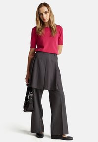Helles pinkes kurzärmliges Sweatshirt, kombiniert mit schiefergrauen, hochtaillierten, plissierten Hosen. Das Modell hält eine schwarze Tasche und trägt flache schwarze Schuhe.