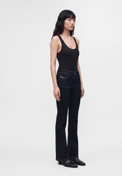 Femme aux longs cheveux noirs portant un haut noir sans manches, un jean bleu foncé et des bottes noires à bout pointu, debout devant un fond uni.
