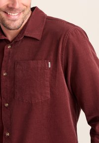 Maroon corduroy shirt featuring a button-up front, a chest pocket, and a small tag. The fabric has a distinct ribbed texture.
