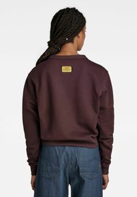 Ciemnobrązowa bluza z prążkowanym dekoltem i mankietami. Z tyłu znajduje się złota metka. Do kompletu luźne, niebieskie spodnie dżinsowe.