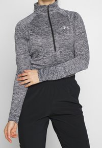 Šedý sportovní top s dlouhým rukávem, vysokým límcem a předním polovičním zipem. Texturovaná látka s raglánovými rukávy, doplněná černými kalhotami.