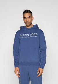 Kék pamut kapucnis sweatshirt, laza szabással. Kanguru zsebbel, húzózsinóros kapucnival és fehér logófelirattal az elején.