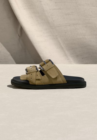Ταμπά σουέντ σανδάλι slide με δύο ασημένιες αγκράφες και μαύρη σόλα σε ανοιχτό μπεζ επιφάνεια και υφασμάτινο φόντο.