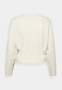 Krämfärgad tröja med en knytning på baksidan, långa ärmar och ribbade muddar. Mjukt, strukturerat material med en kortad design.