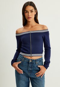 Marinblå off-shoulder crop top med en silvrig dragkedja och vit randig detaljer vid halsringningen och nederkanten. Matchas med högmidjade jeans.