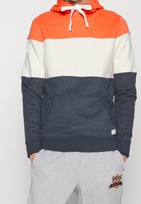 Felpa con cappuccio a blocchi di colore, con parte superiore arancione, parte centrale crema e parte inferiore blu navy. Presenta una tasca frontale e cappuccio con cordino. Tessuti morbidi.