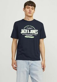 Tamsiai mėlynas medvilninis marškinėliai su dideliu baltu ir žaliu grafinio teksto dizainu, kuris ant priekio rašo "JACK & JONES". Trumpomis rankovėmis.